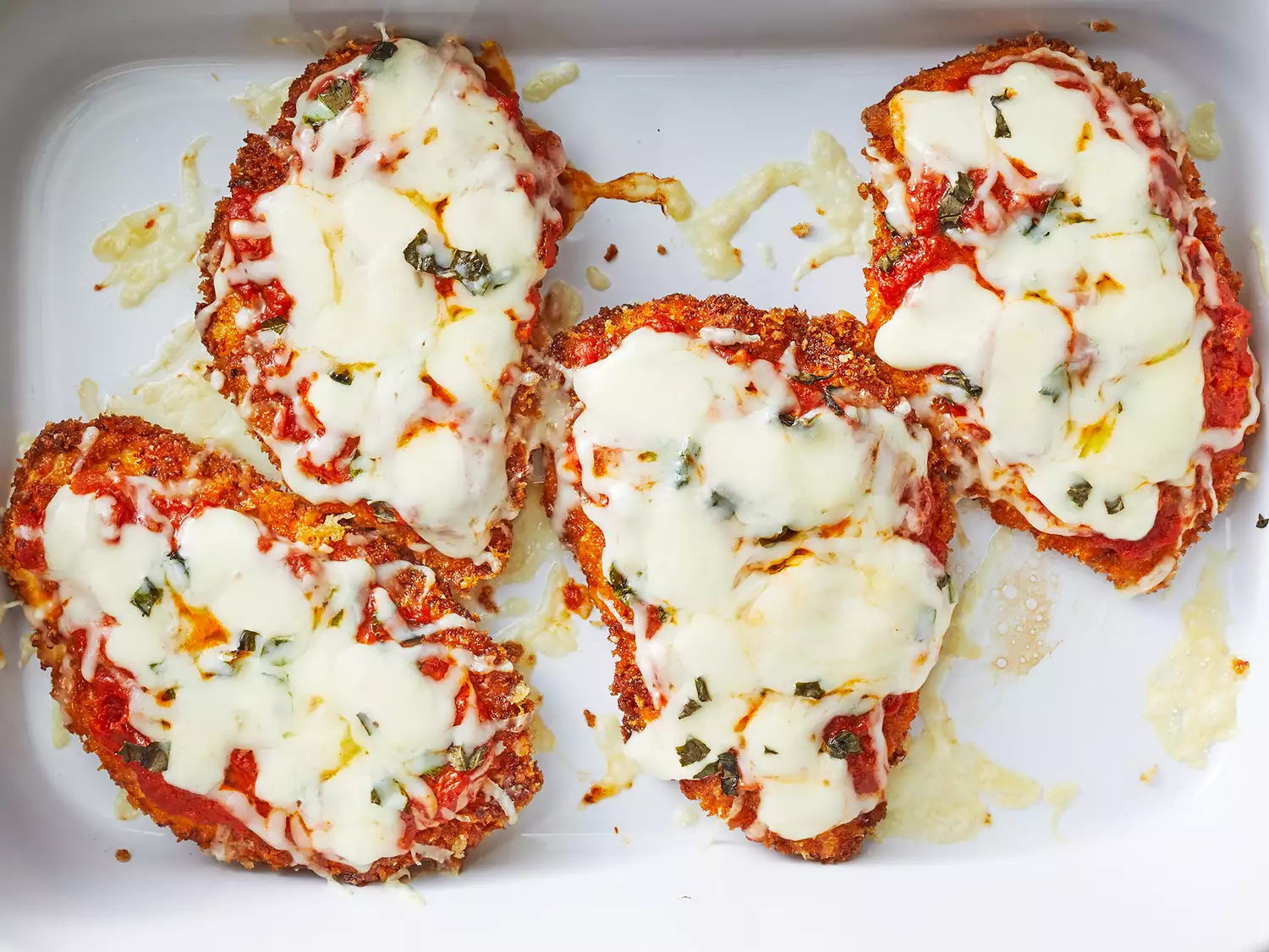 Four fully-cooked and browned chicken parmesan with melted white cheese and chopped basil all in a white cooking dish. 
            Image found on https://www.allrecipes.com/recipe/223042/chicken-parmesan/